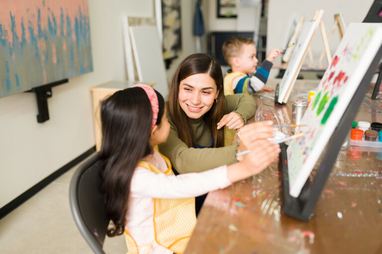 Hispanic female instructor smiling and teaching a little girl how to paint on a canvas during a painting class for children